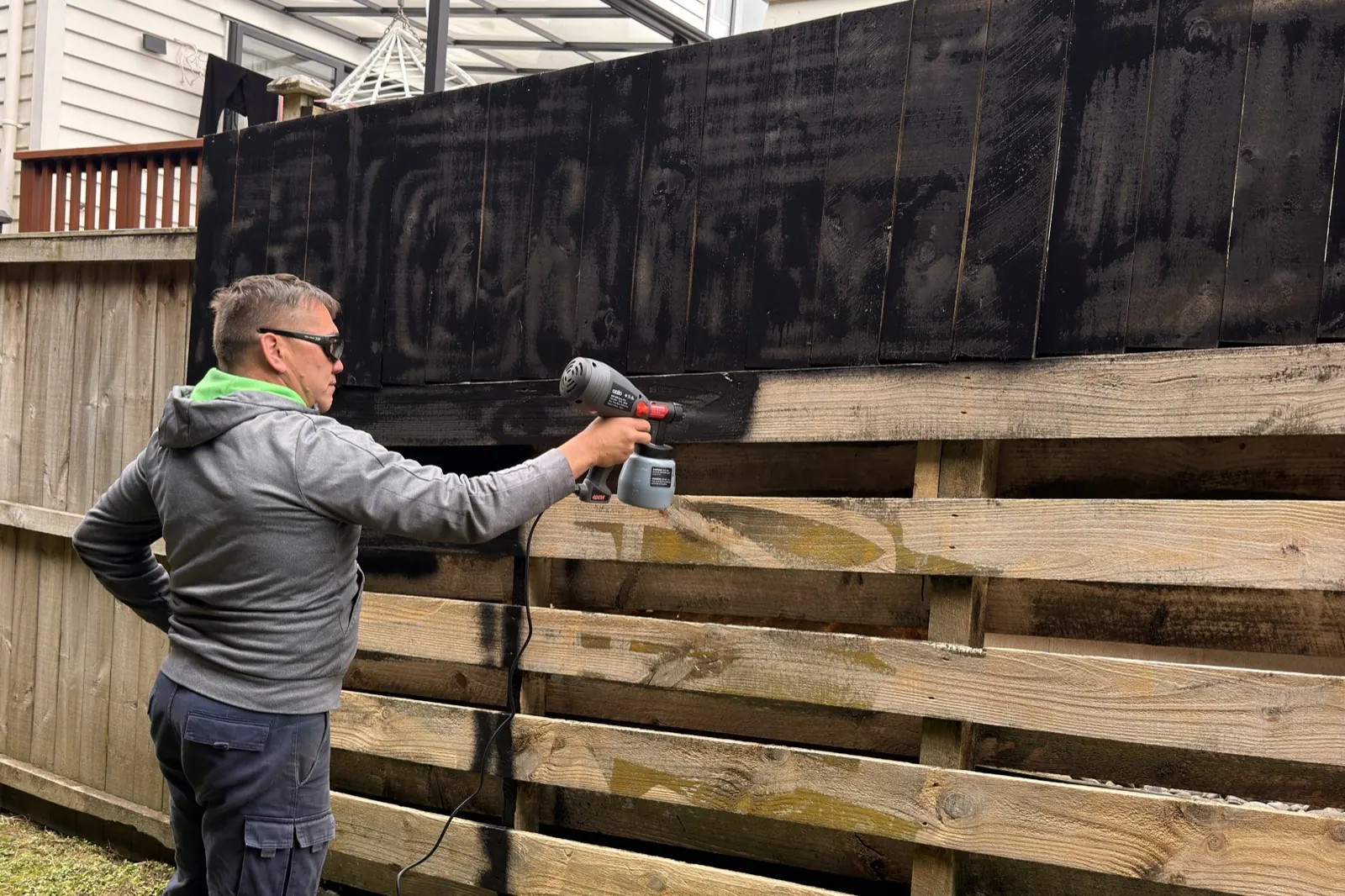 Rinat spraying the top section of the fence black with an electric spray gun, the lower rails still bare timber