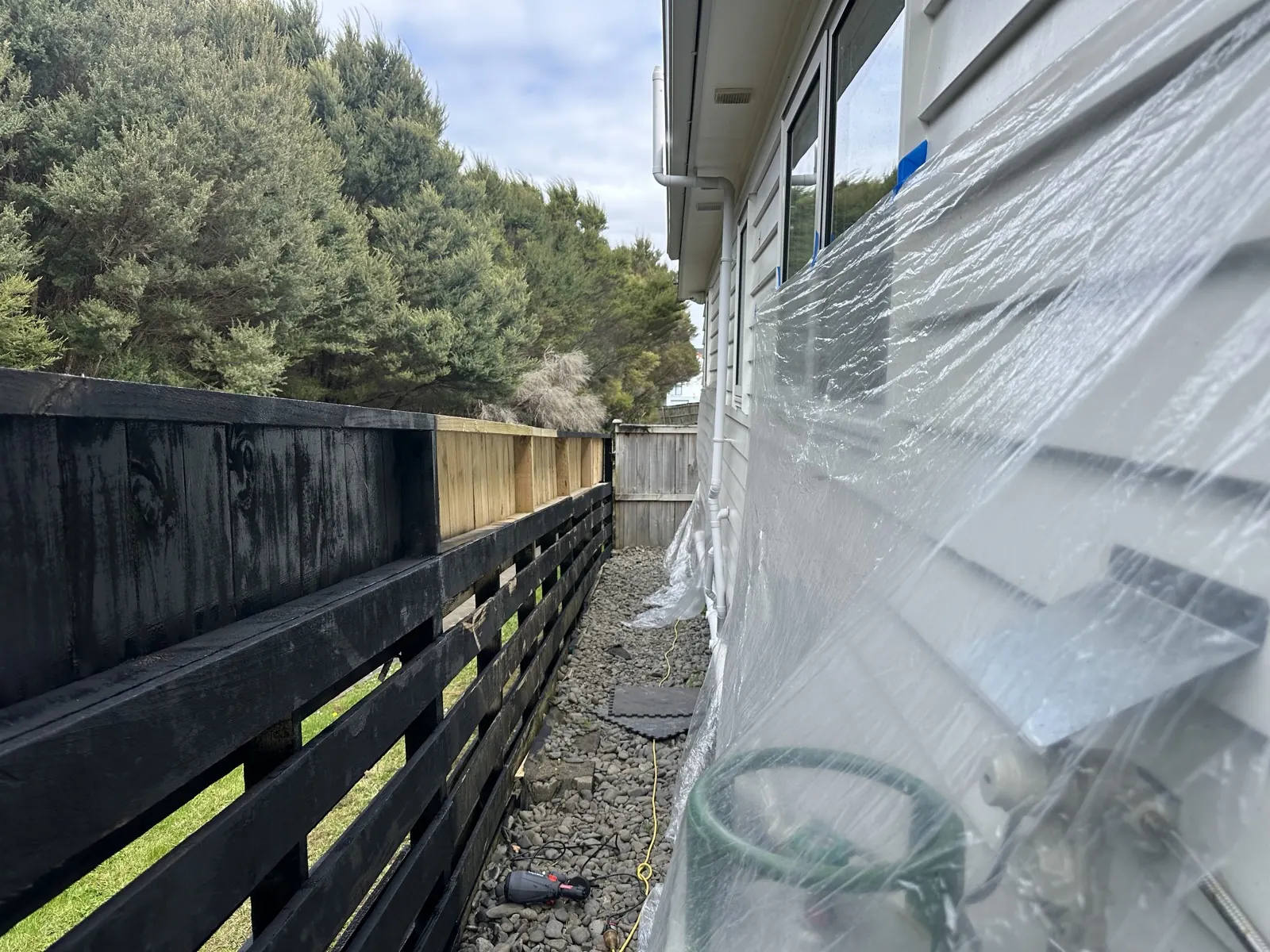 The house wall and windows masked off with plastic sheeting while the fence is sprayed black