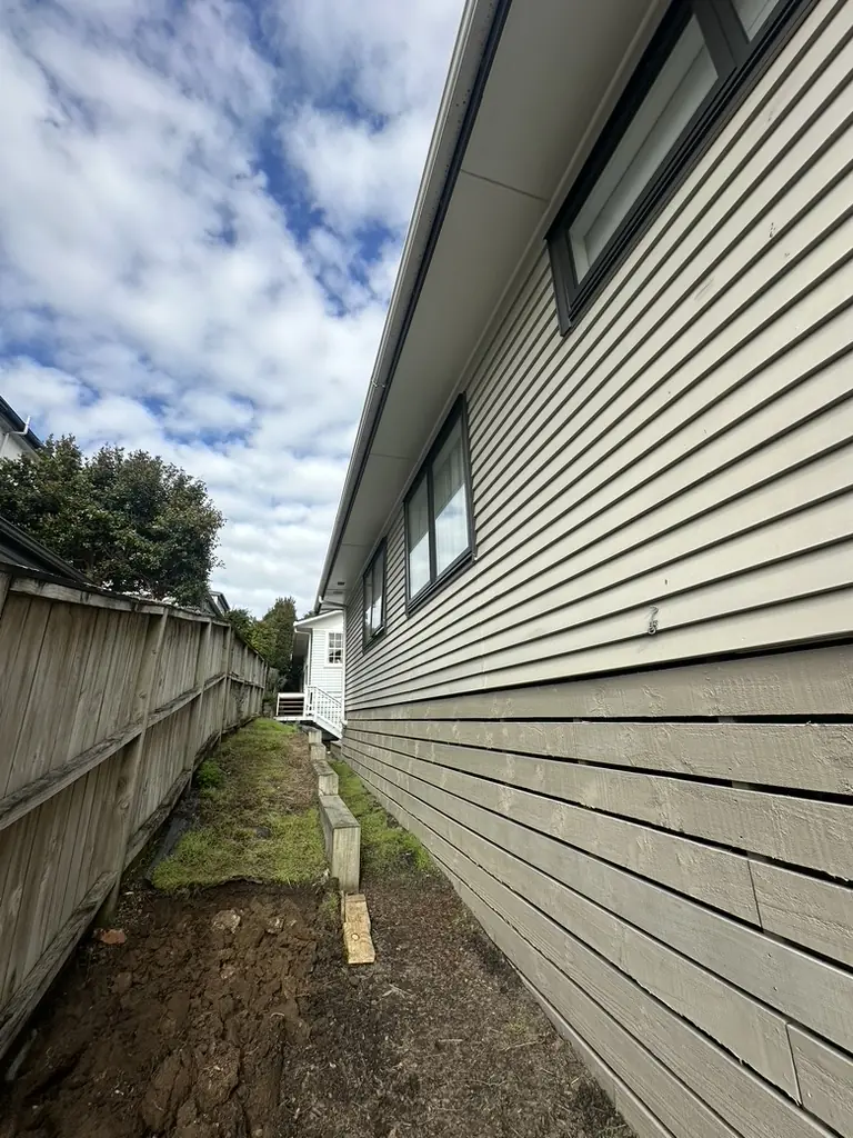 Narrow side passage between a weatherboard house in Howick and the boundary fence, with the gutter running high along the eaves