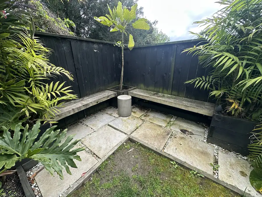 Quiet seating corner in a Pakuranga back yard before water blasting, with moss greening the pavers, the timber bench and the dark fence
